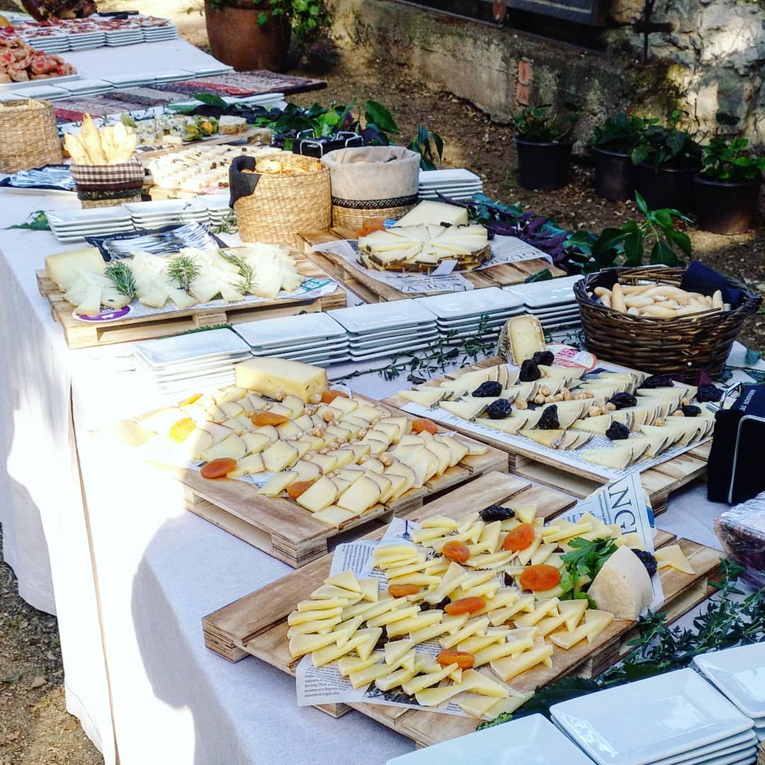 A rustic outdoor market stall displays an array of fresh produce including vibrant tomatoes, leafy greens, and a variety of olives, set against the backdrop of a picturesque rural Italian village.