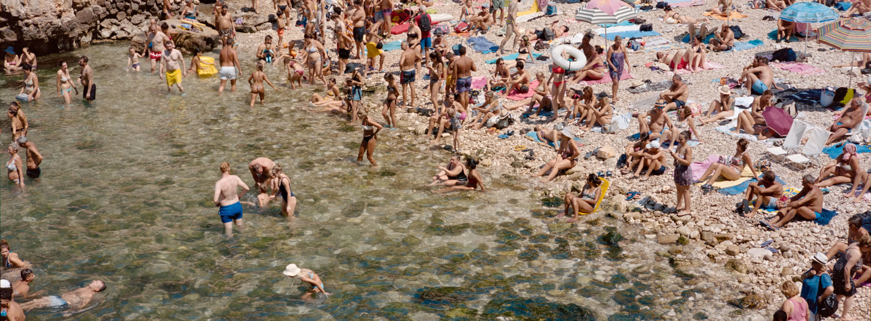 Italian summer beach scene captured on film with a Hasselblad XPan, featuring a bustling crowd of people swimming and sunbathing along the picturesque coastline, showcasing the camera's wide-format cinematic aesthetic for documentary-style photography