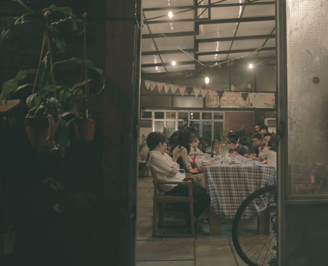 A group of people sits around a table covered with a checkered tablecloth in a dimly lit indoor space. The table is set with plates and glasses. String lights hang from the ceiling, which has a translucent or glass roof structure. A bicycle wheel is visible near the table, and potted plants hang from the left side. The scene is captured from a doorway or passageway, with dark walls and flooring visible in the foreground.