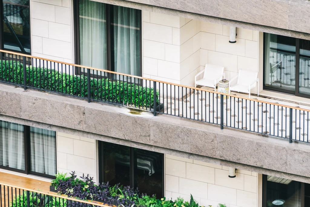 Modern balcony with gray tile flooring, glass railing overlooking greenery. Minimalist white sofas and two potted plants create a serene atmosphere.