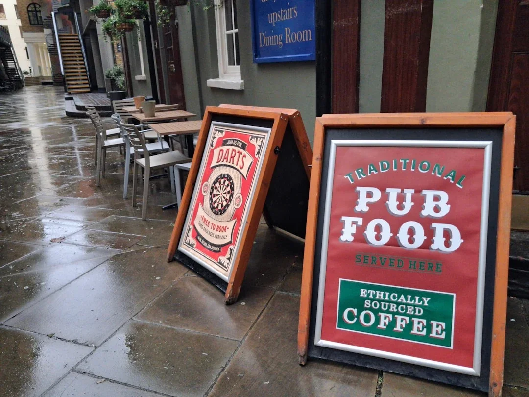 Two a-frame signs outside a pub on a wet street.