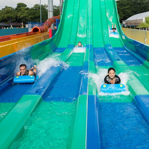 People slide down colorful water slides on mats, splashing into a pool, with greenery and a covered structure in the background.