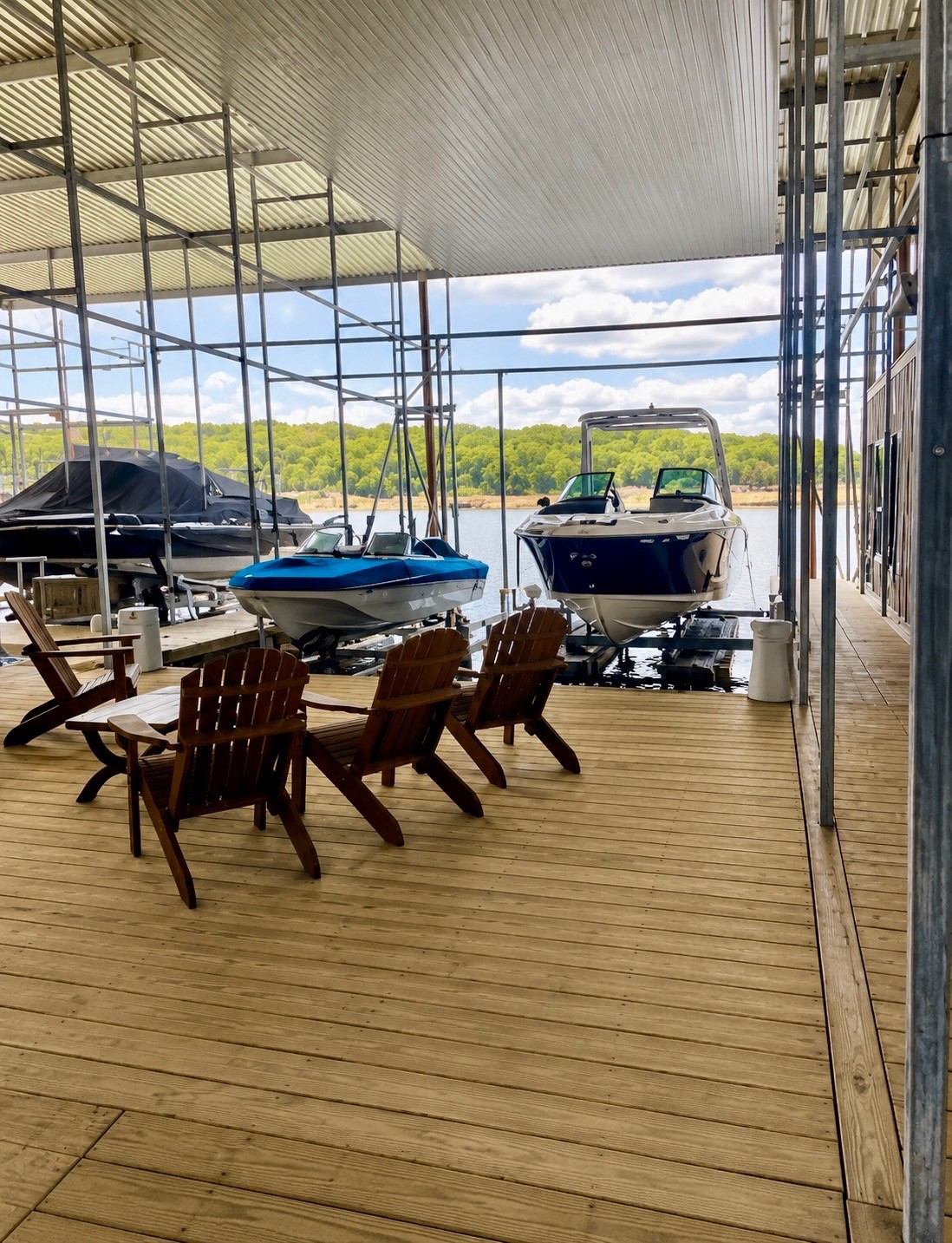 A covered dock with wooden chairs overlooks motorboats secured in slips, surrounded by a scenic view of a calm lake and tree-lined shore under a partly cloudy sky.