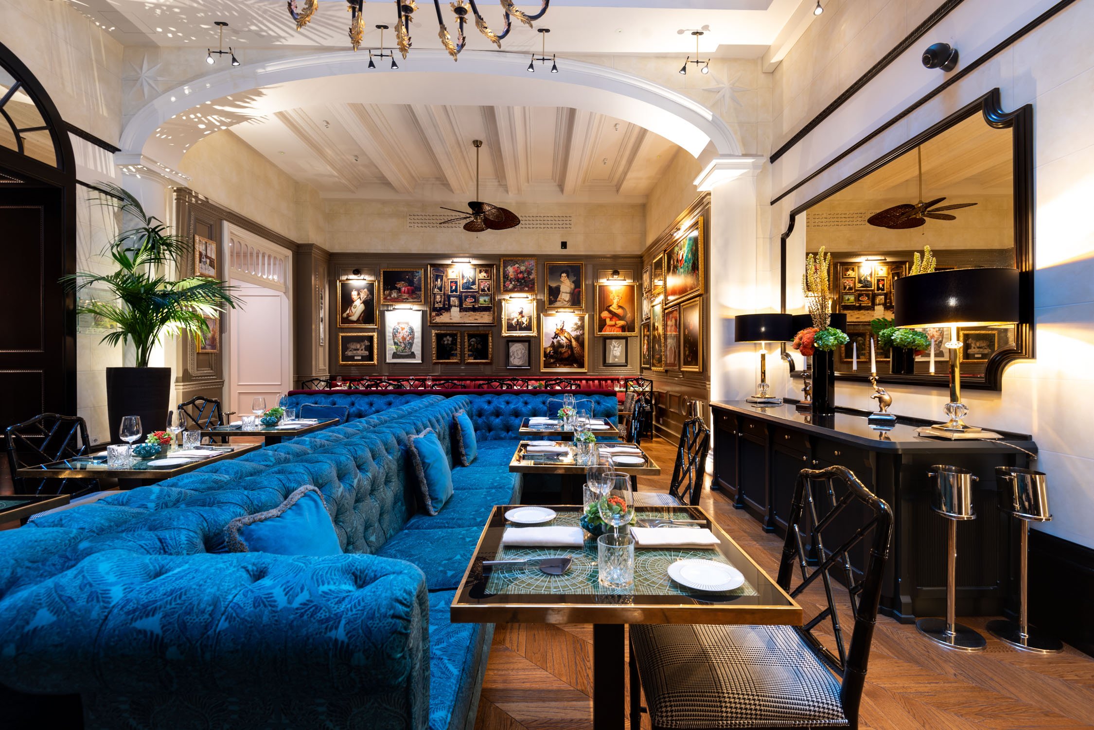 A dimly lit lounge with plush blue velvet booths, a long bar with brass accents, and warm overhead lighting.
