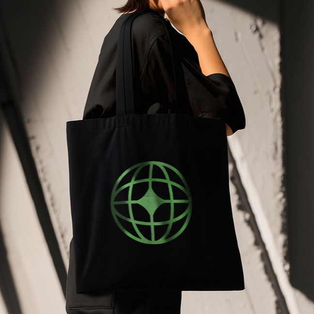 Person wearing black clothing carrying a black tote bag with a green globe-like geometric symbol, standing against a concrete wall in sunlight and shadow.