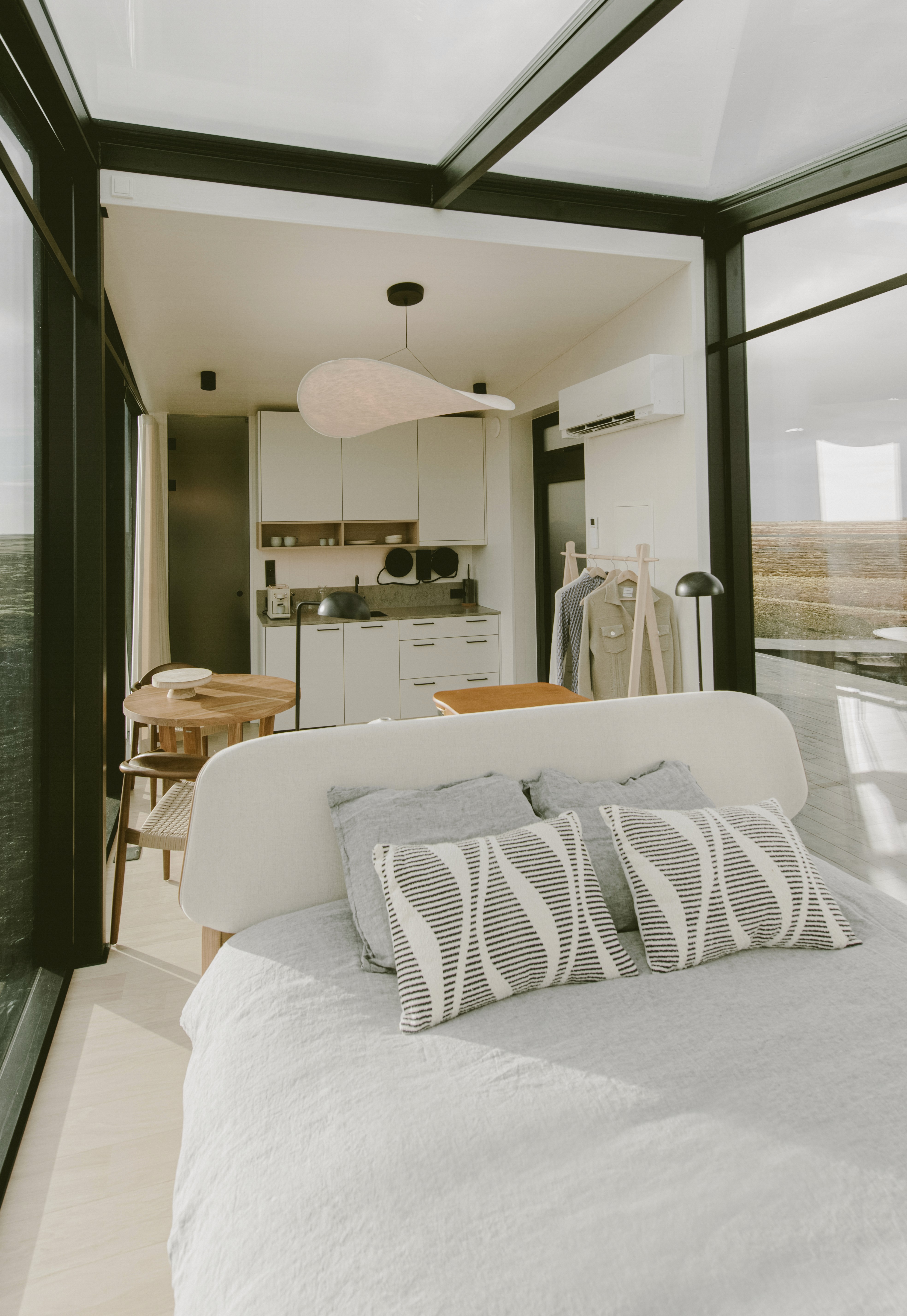 Bright open-plan cabin interior featuring a minimalist bedroom, compact kitchen and dining area, with floor-to-ceiling glass walls and contemporary design overlooking a vast natural landscape.