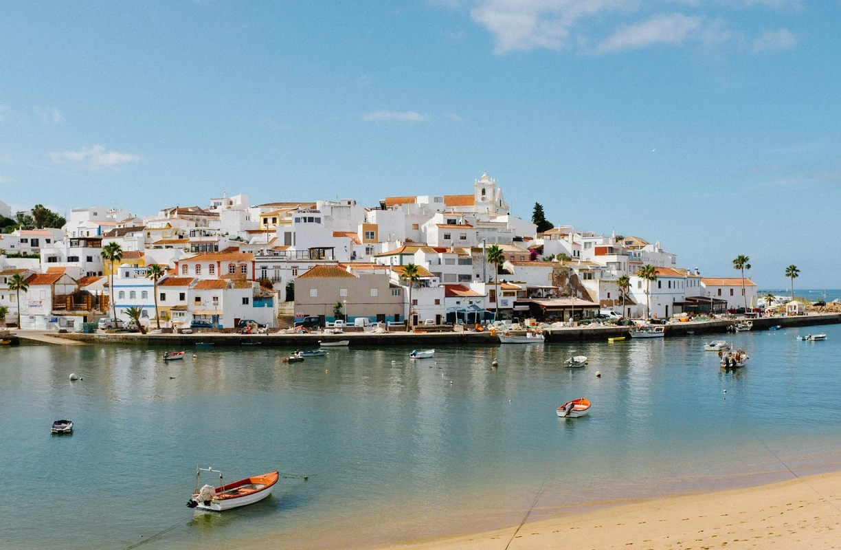 Scenic view of a charming coastal town in portugal with whitewashed buildings, colorful boats floating on calm waters, and a sandy beach under a clear blue sky