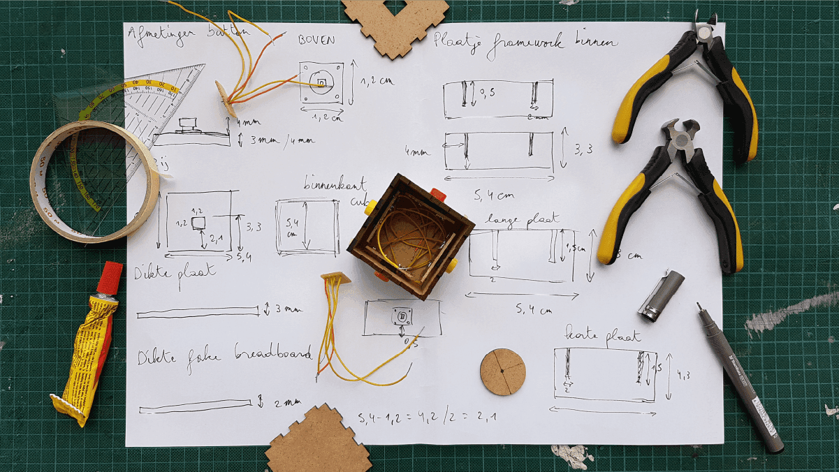 Prototype sketches and measurements on paper with a small cardboard model, tools, and craft supplies on a cutting mat