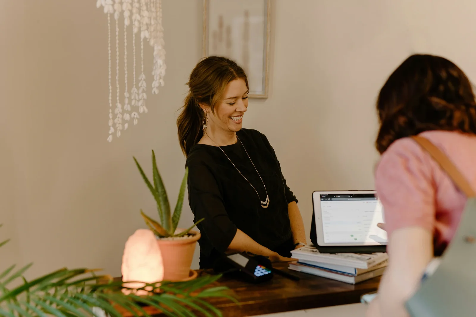 Woman in black shirt smiles while assisting customer at desk with laptop and plant in modern office setting.