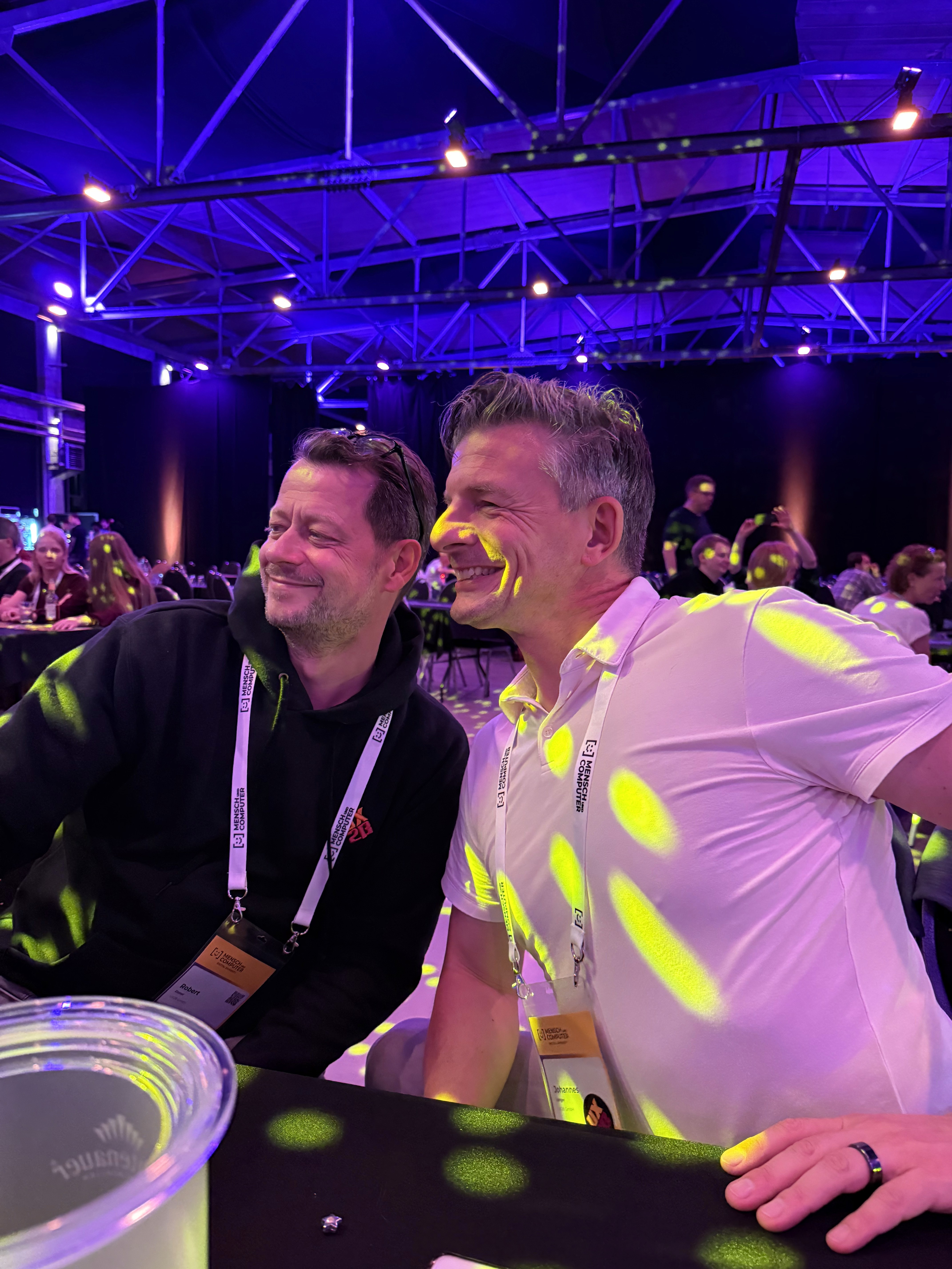 Zwei Männer mit Namensschildern sitzen an einem Tisch in einem Veranstaltungsraum mit violetter Beleuchtung