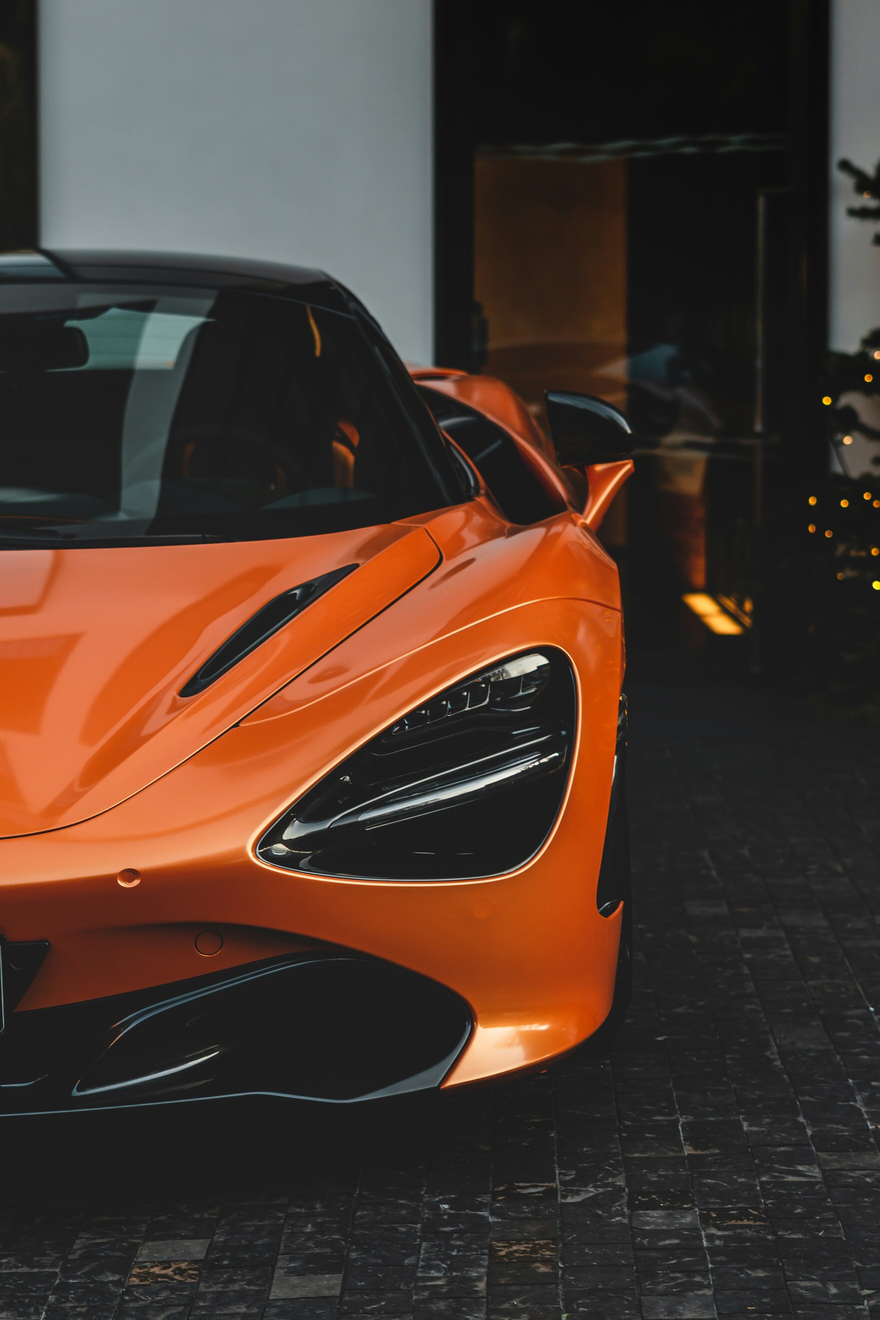 A sleek, orange luxury sports car is parked on a dark cobblestone driveway, showcasing its aerodynamic design and modern headlights.