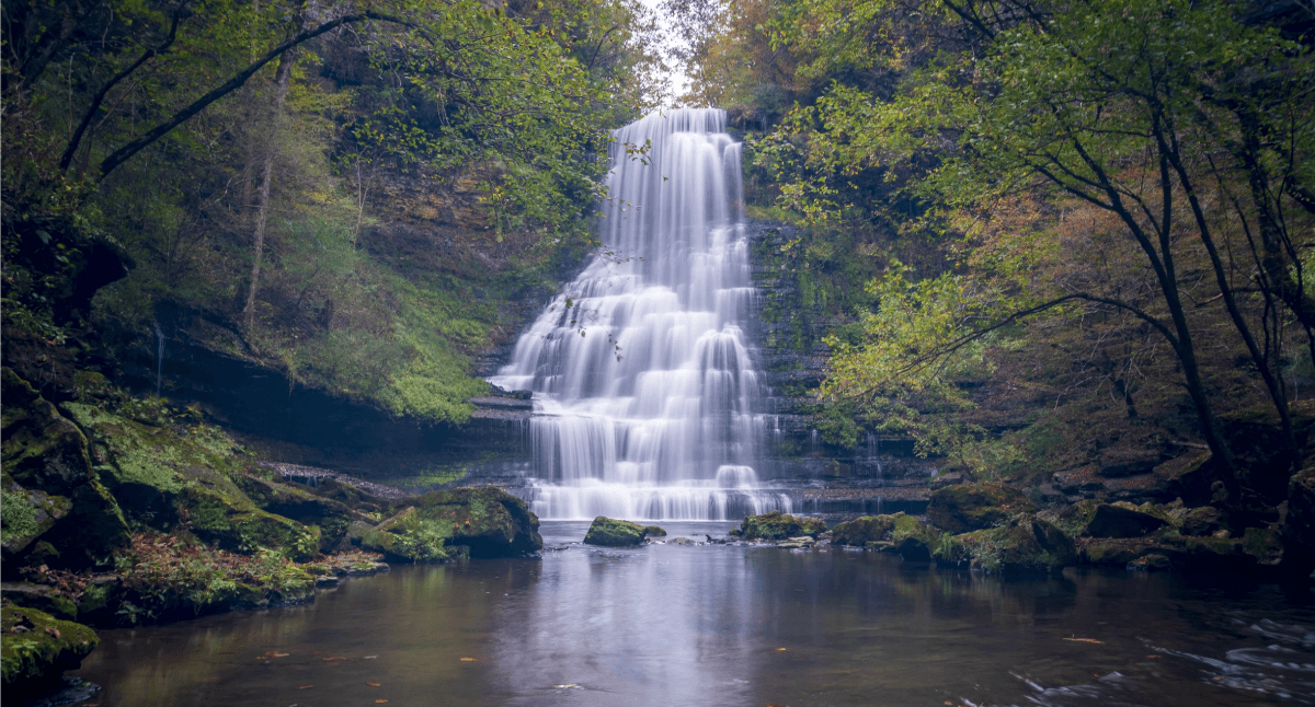 A rocky mountain landscape with a cascading waterfall flowing down its cliffs surrounded by greenery