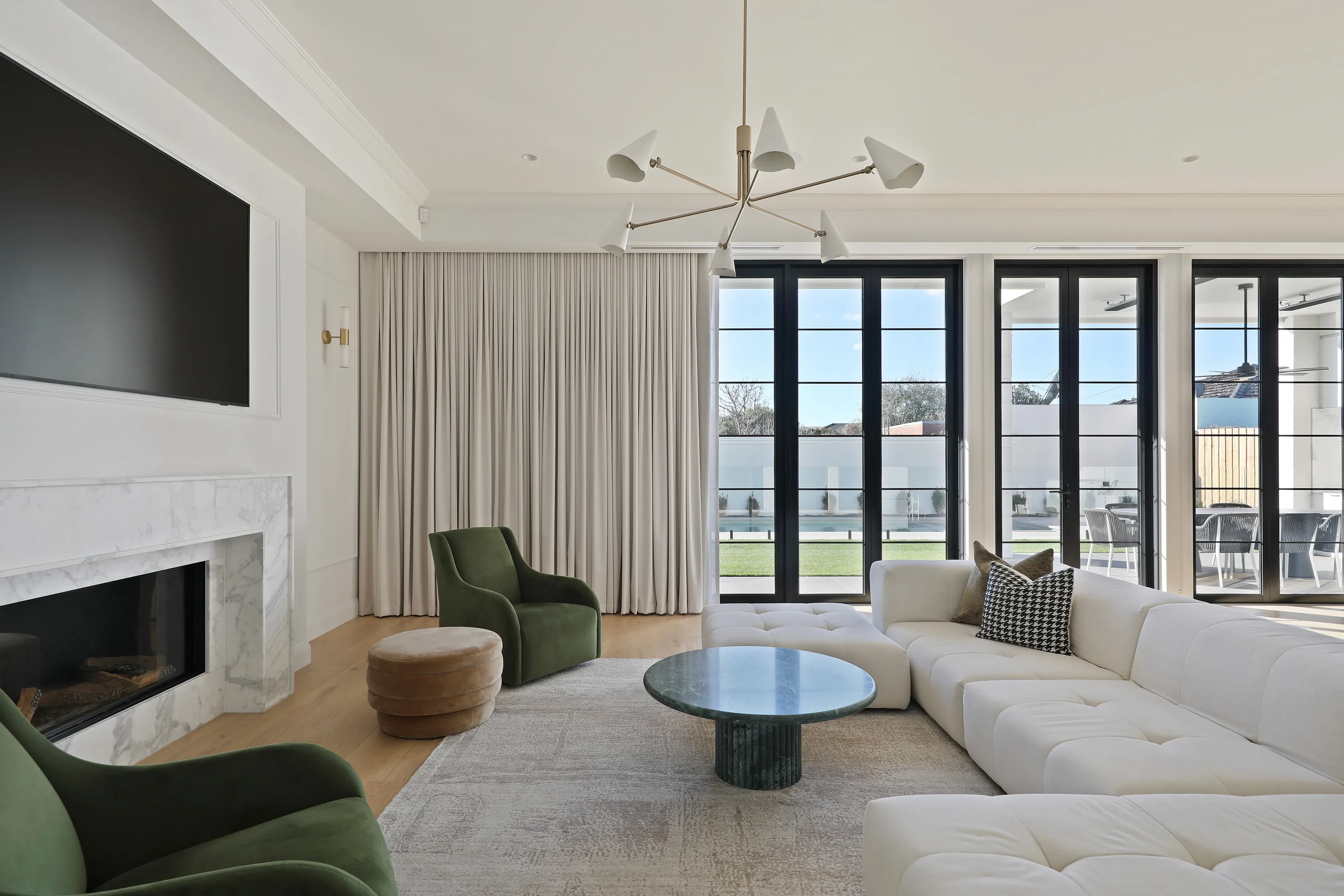 Living room with a black-panelled TV wall over a white marble fireplace, a white modular sofa, olive velvet armchairs, a green marble coffee table and a wall of black-framed doors to the pool