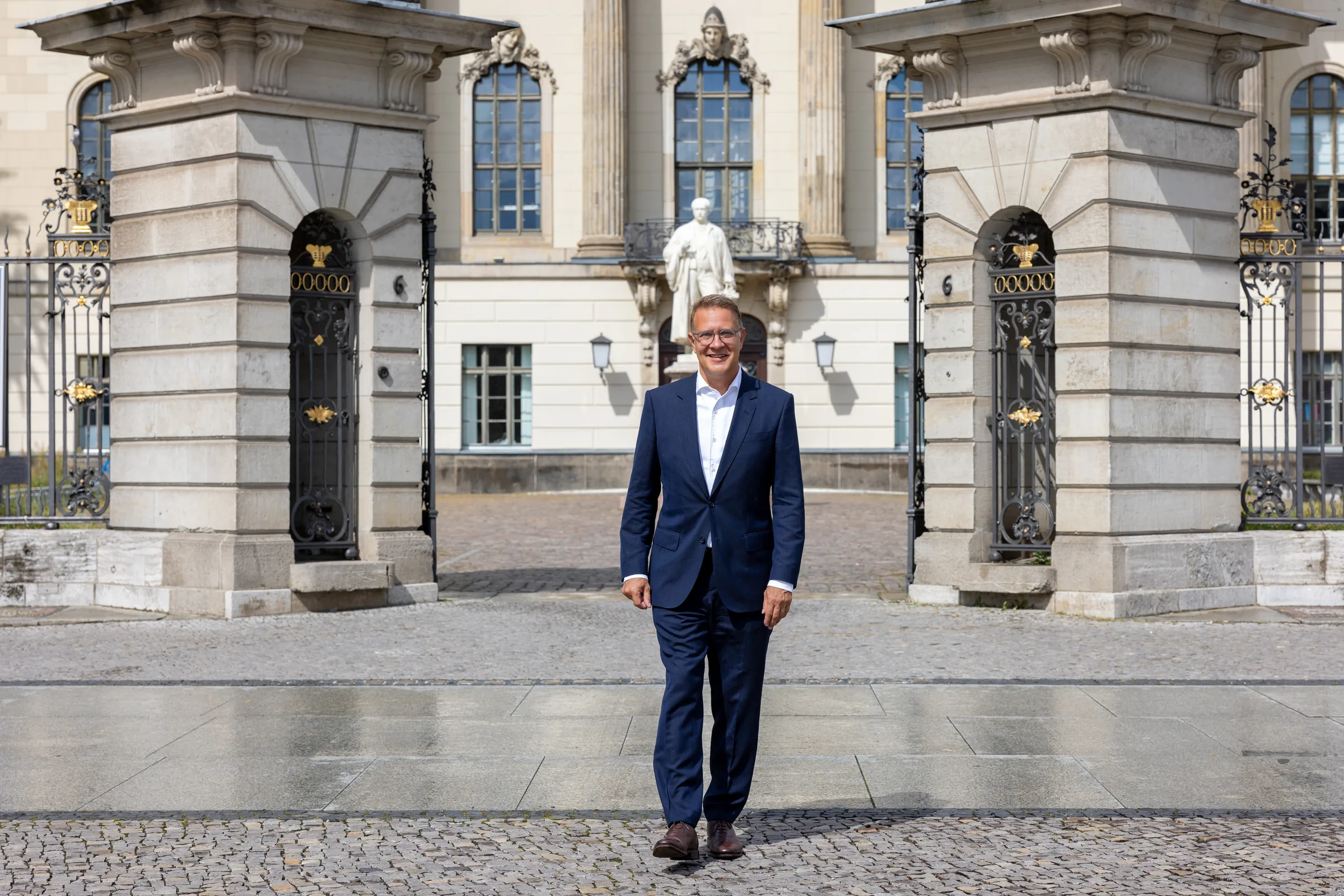 Adrian Grasse geht lächelnd durch ein imposantes, schmiedeeisernes Tor vor einem historischen Berliner Gebäude mit Säulen und Statuen.