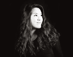 A black and white portrait of a woman with long, wavy hair. She is looking off to her right, against a dark background. She wears a dark top. The image is in black and white.