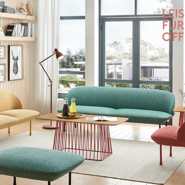 A modern living room featuring a curved teal sofa with a geometric pattern, a matching ottoman, and a yellow loveseat. A rectangular wooden coffee table with a red wire cage base sits on a light gray rug, accented by a red floor lamp and large windows in the background.
