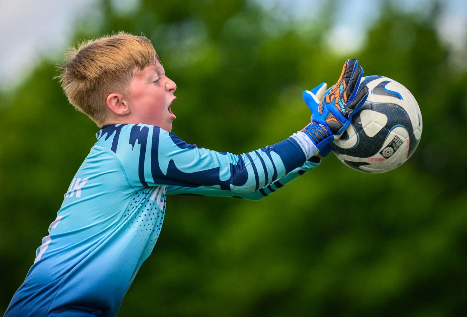 Side view of a young male youth goalkeeper in a blue uniform reaching upward and shouting as he goes for a soccer ball outdoors with greenery in the background.