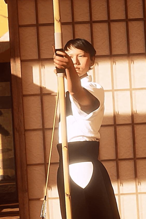 Kyudo archer holds a long bow in warm sunlight against a shoji screen.