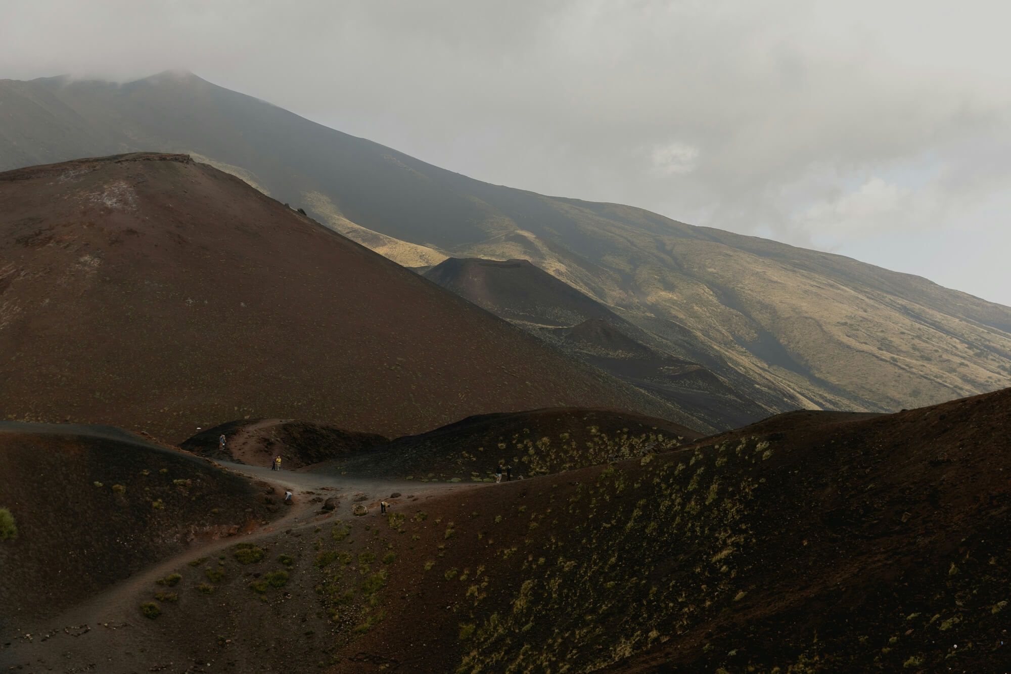 Dark volcanic hills under a cloudy sky, with a winding path cutting through. Sparse vegetation hints at a rugged, desolate landscape.
