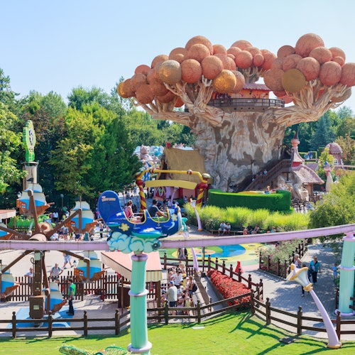 פארק שעשועים עם מתקנים צבעוניים, מבנה עצים גדול ואנשים שנהנים מאטרקציות שונות תחת שמיים כחולים ובהירים.