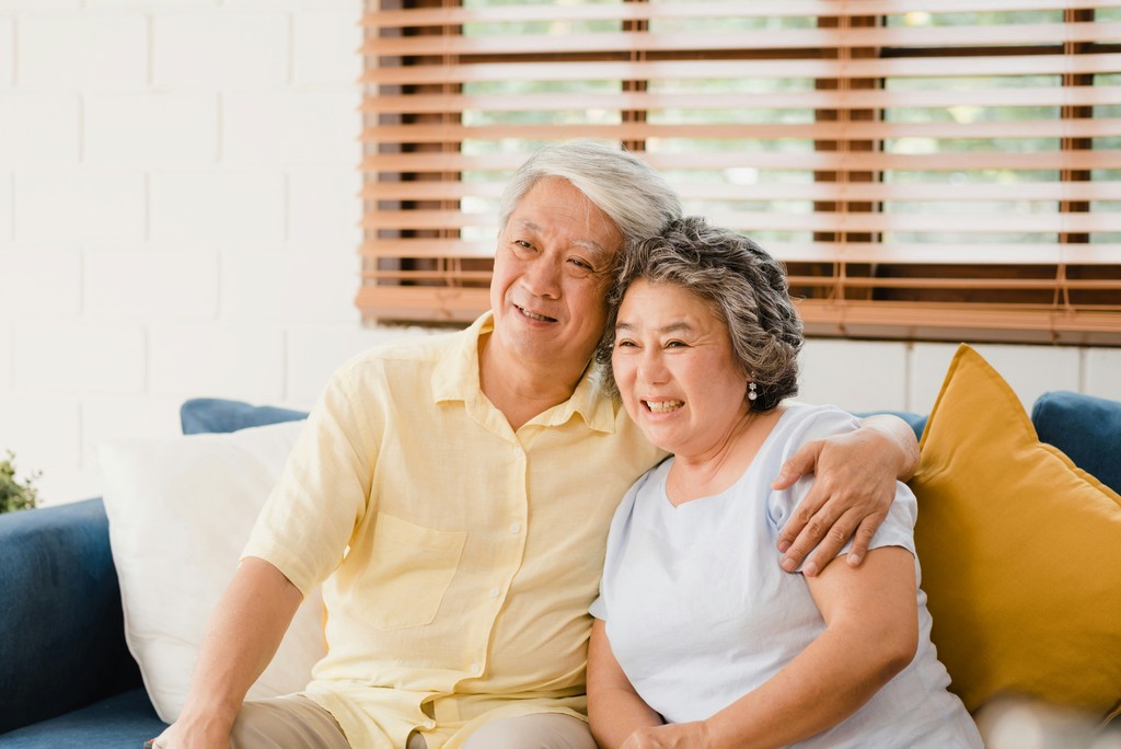 A happy elderly couple sits closely together on a cozy sofa, smiling and embracing, conveying warmth, love, and companionship in a comfortable home setting.