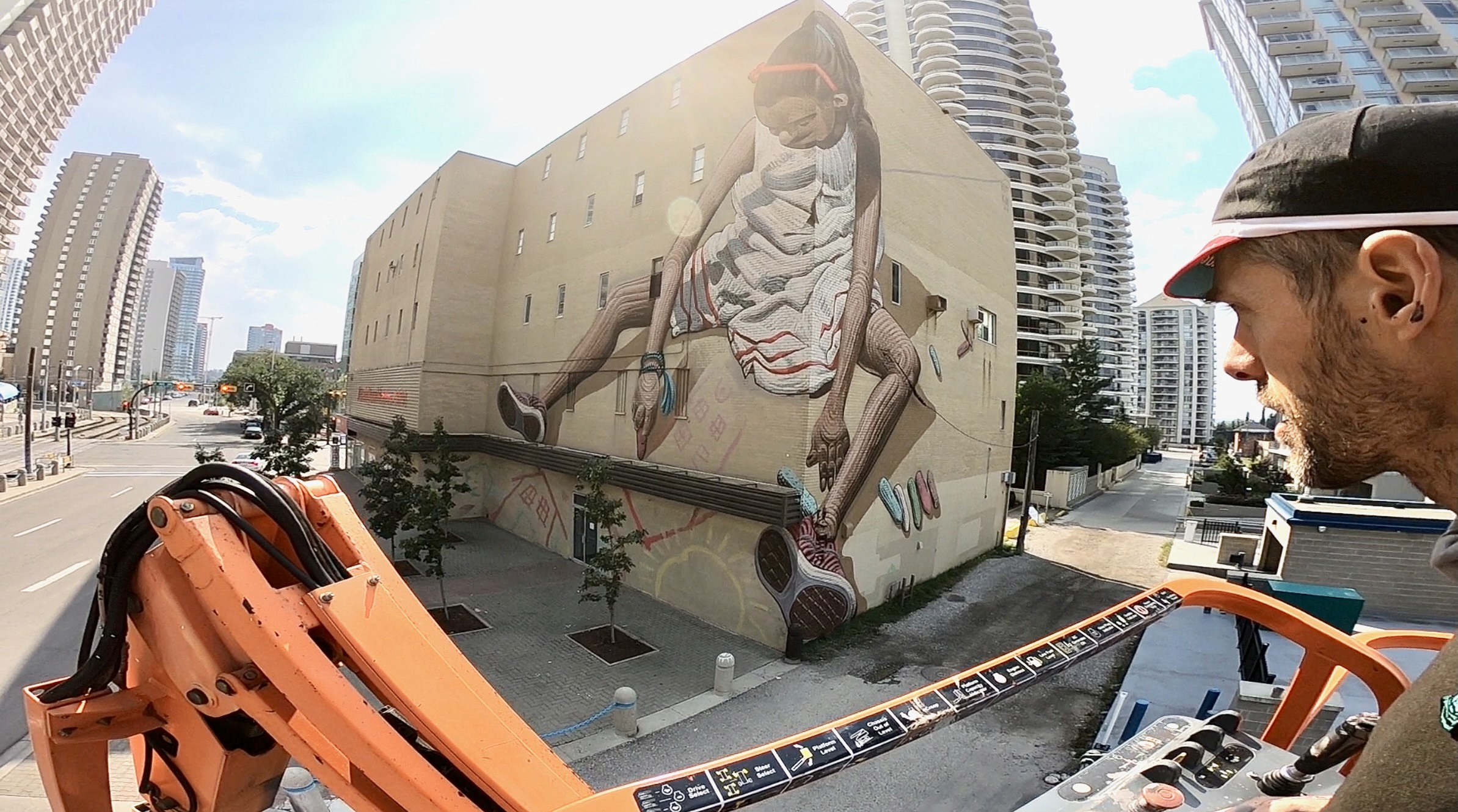 From a lift basket, a man looks back at a girl painted across a tall beige building.