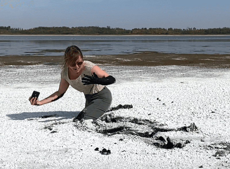 A video still of a woman sinking into white sand, looking with horror at her arm that's covered in tar-like sludge.