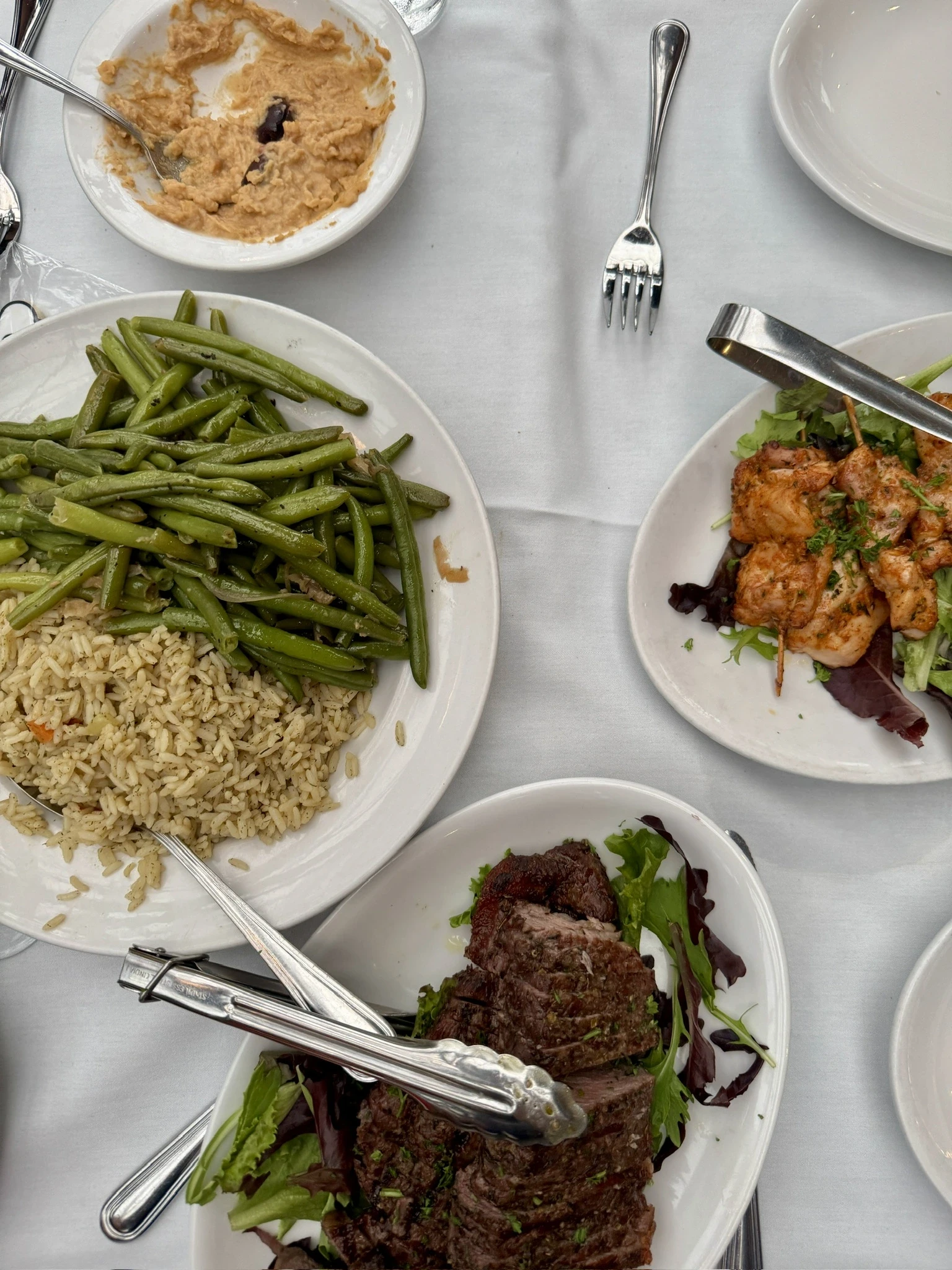 Greek meal on a white table cloth in downtown Raleigh, NC with green beans, hummus, grilled chicken skewers, and steak