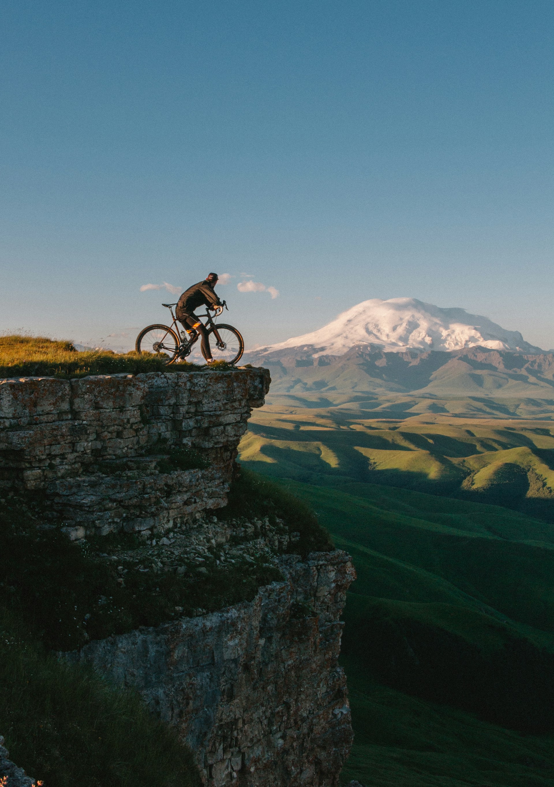 A cyclist standing with their bike on a grassy cliff edge, looking out toward a massive snow-capped mountain peak.
