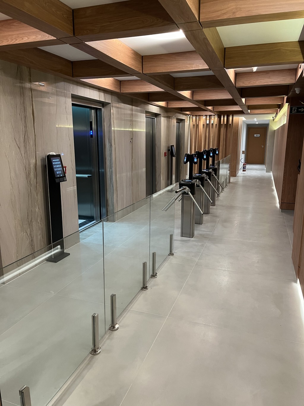 Vista interna do saguão de elevadores do Trade Center Dom Campelo mostrando corredor moderno com acabamento em mármore claro nas paredes, detalhes em madeira no teto, sistema de catracas eletrônicas com biometria e guarda-corpos de vidro, iluminação LED integrada criando ambiente corporativo sofisticado.