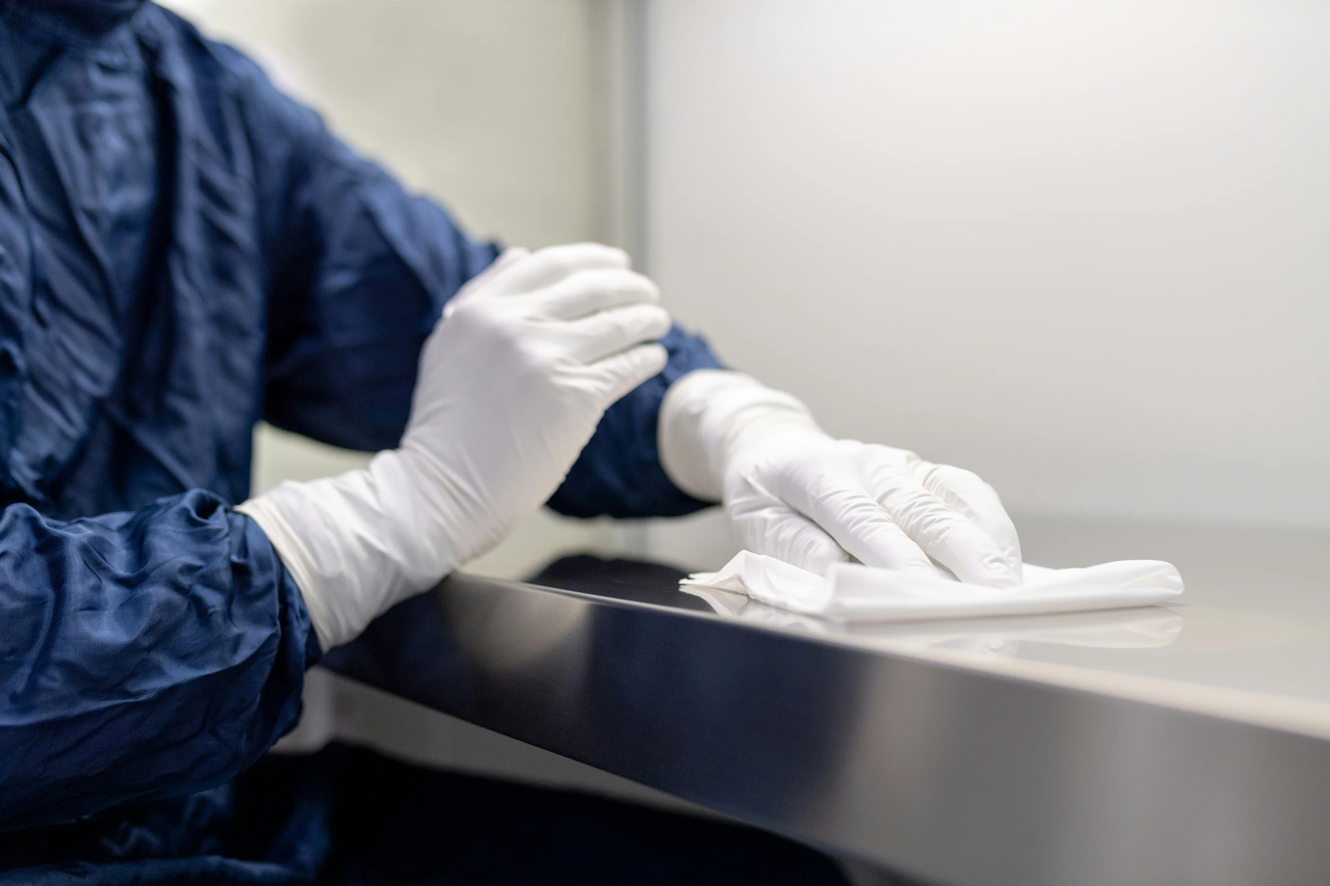 Nahaufnahme einer Person in blauer Schutzkleidung und weißen Schutzhandschuhen, die mit einem weißen Einwegtuch sorgfältig eine metallische Tischoberfläche abwischt.