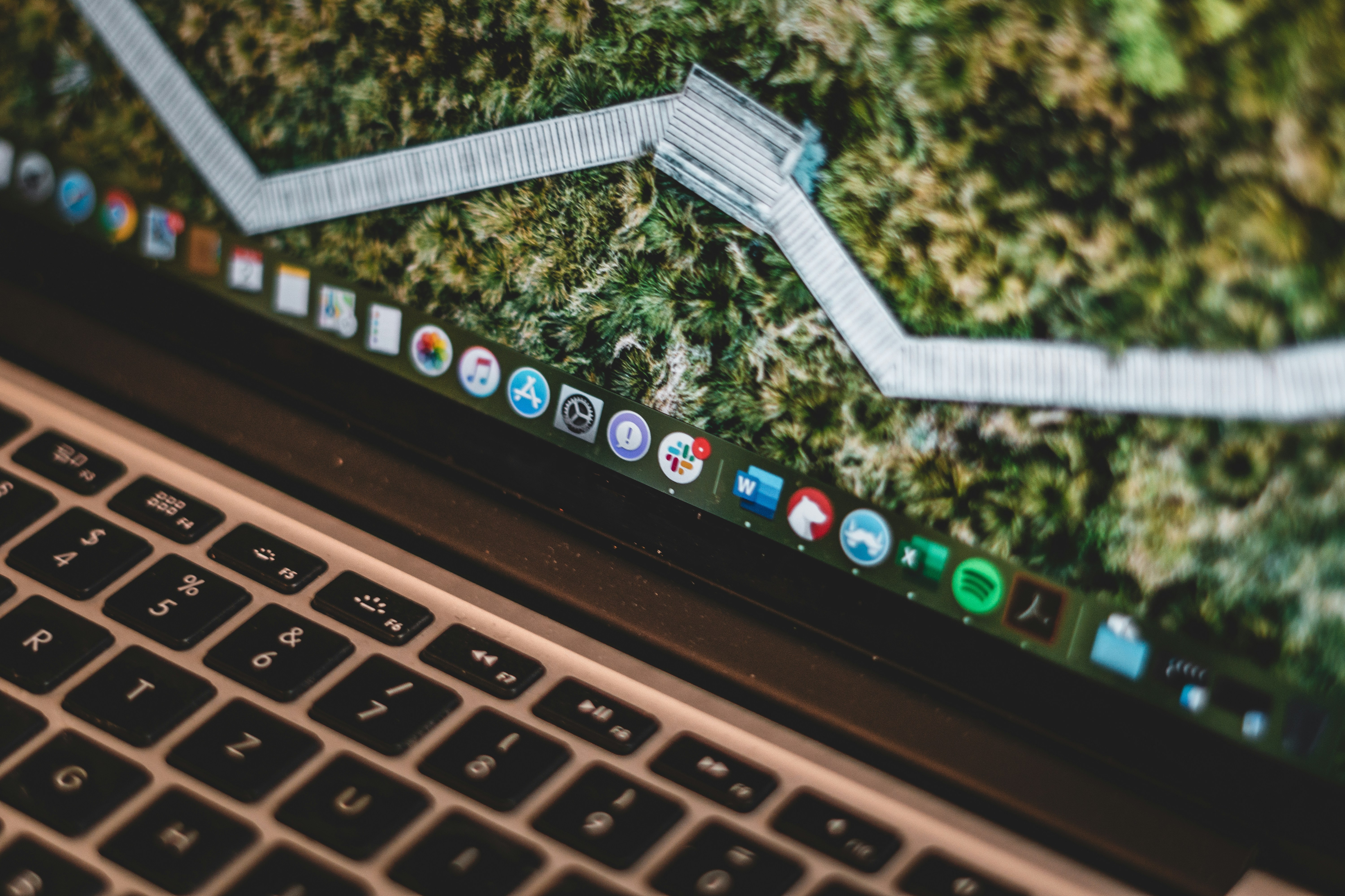 Laptop keyboard with dock bar showing multiple application icons against a blurred green foliage background