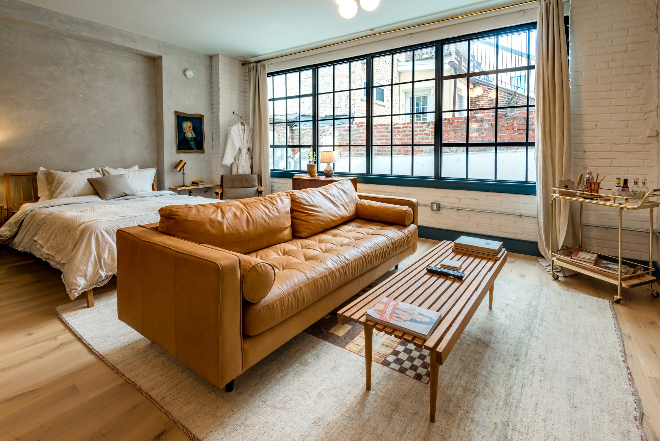 A modern studio apartment with a leather sofa and bed in front of large industrial windows overlooking a city street.