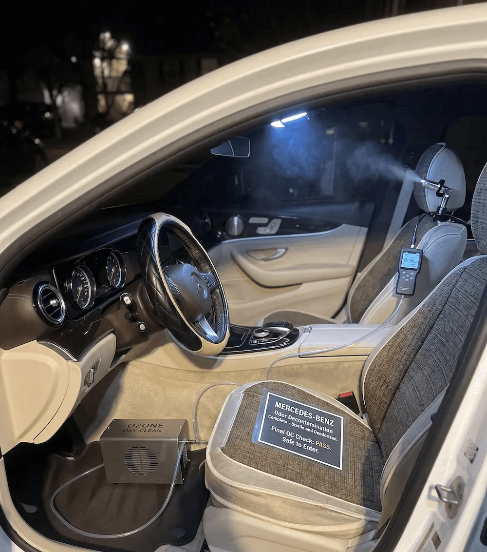 A professional nighttime shot of a Mercedes-Benz interior undergoing odor decontamination. The beige and brown cabin is illuminated by a bright interior light, showing an ozone machine on the driver-side floor connected by a clear tube to a misting nozzle mounted on the passenger headrest. A digital sensor displays real-time data, and a formal "Final QC Check: PASS" certificate is displayed on the passenger seat, emphasizing a clean and safe environment.