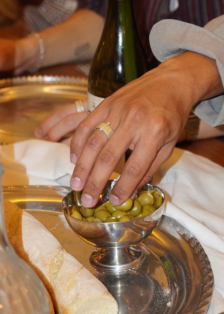 Person holding a silver bowl of olives with bread and a bottle in the background. With silver rings.