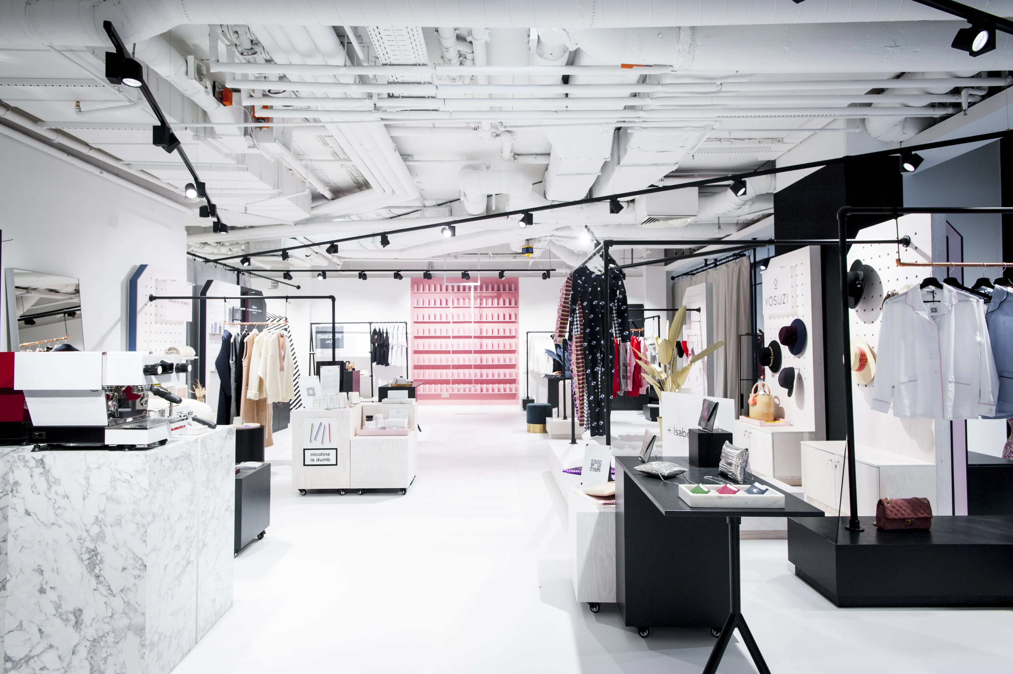 Retail floor at FREIRAUM SHOP with black track lighting, marble and white display counters, clothing rails, a pink spray-can shelf wall in the back and bright white flooring.