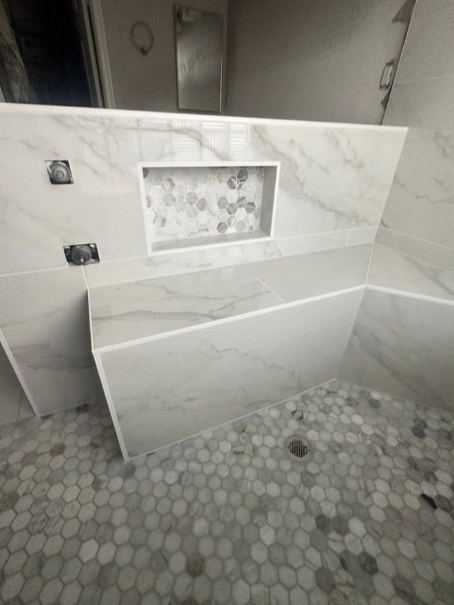 Bathroom shower showcasing large format white marble-look wall tiles, an angled half-wall, a built-in shower niche with a decorative hexagonal mosaic, and a matching hexagonal mosaic shower floor.