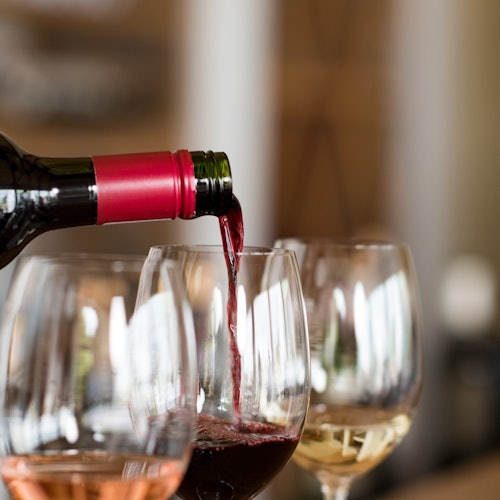 Láhev s červeným uzávěrem nalévá červené víno do sklenice, přičemž dvě další sklenice naplněné bílým a růžovým vínem stojí poblíž.