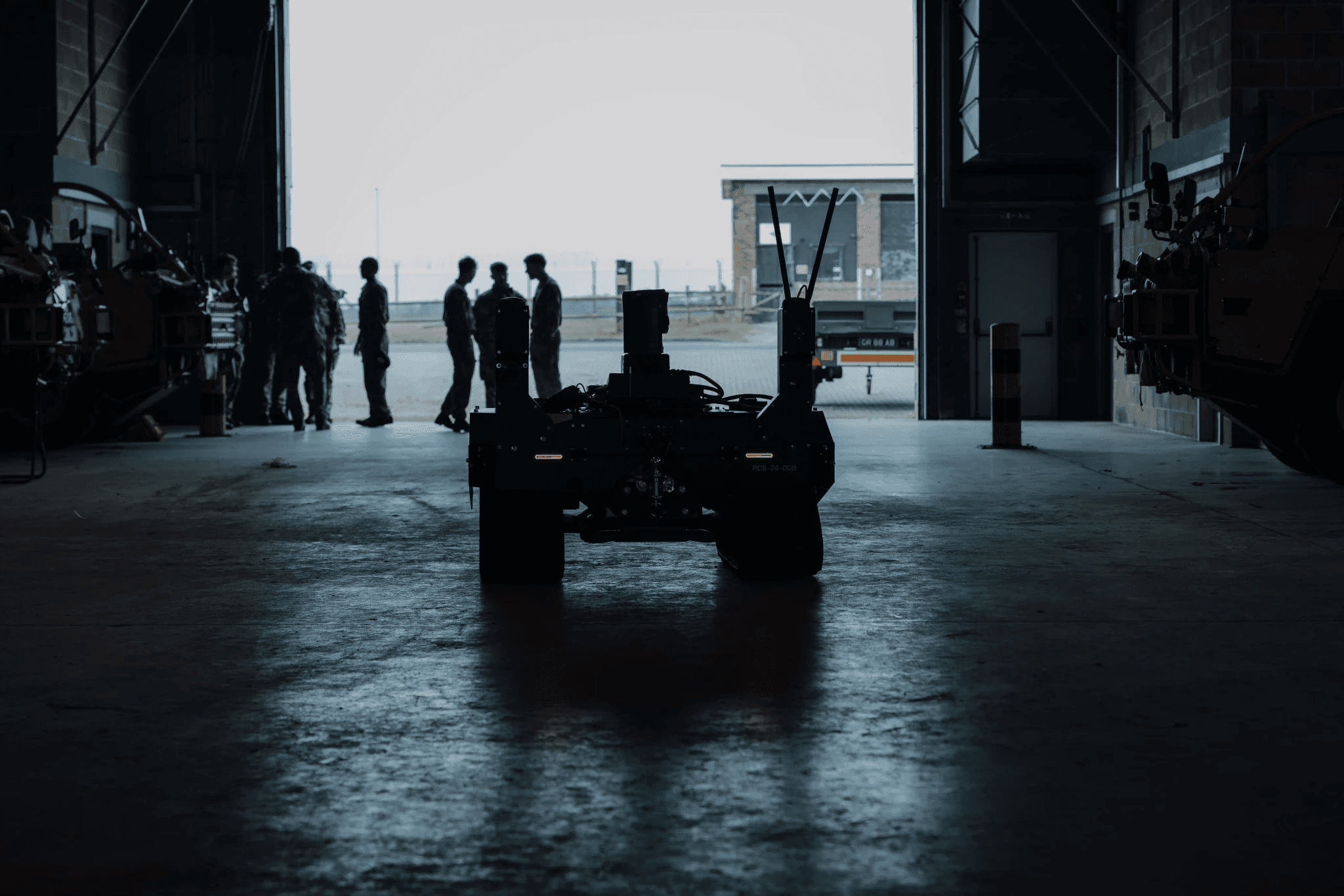 A silhouette of military personnel and a vehicle inside a large, dimly lit hangar, with light streaming in from an open entrance, highlighting the figures and creating dramatic shadows.