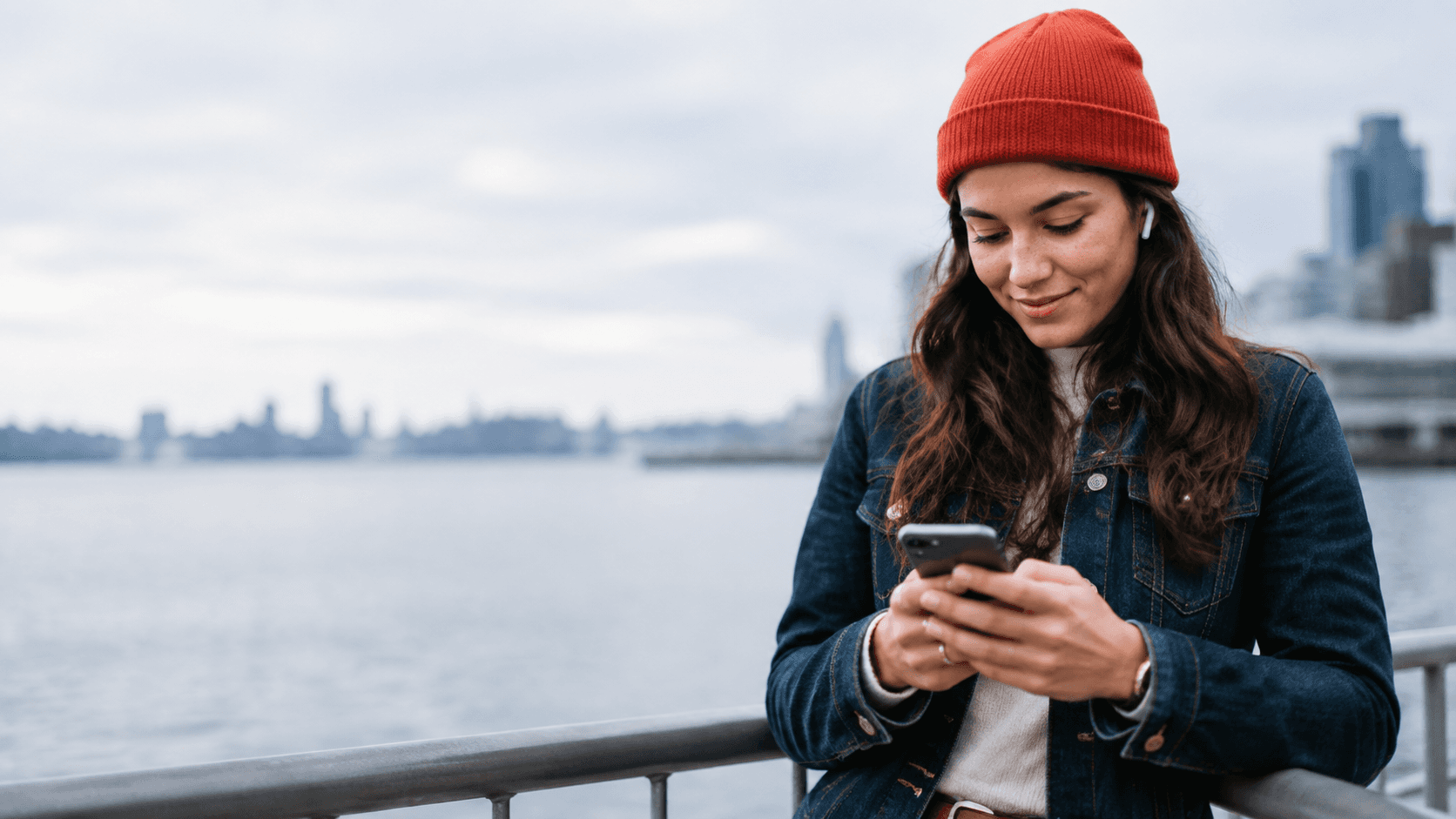 A young woman uses her smartphone outdoors beside an urban waterfront.