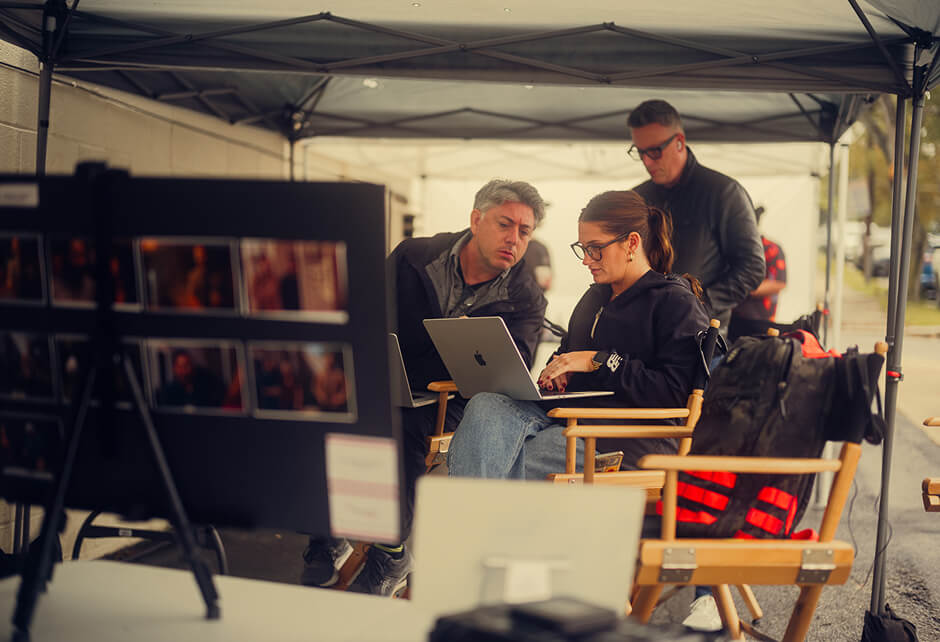 Three people work under a canopy; two are seated looking at a laptop, while one stands behind them. A storyboard and equipment are visible in the foreground.
