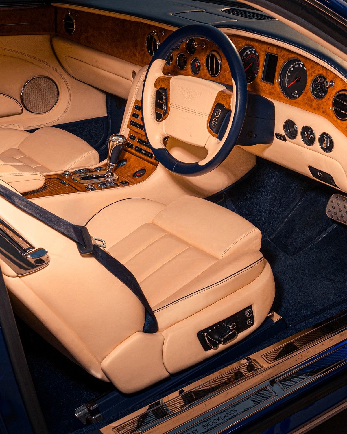 Interior of a Moroccan Blue Bentley Brooklands showing the magnolia leather driver's seat, navy dashboard and carpet, burr walnut veneer, and the winged-B steering wheel, with "Bentley Brooklands" engraved on the door sill plate.