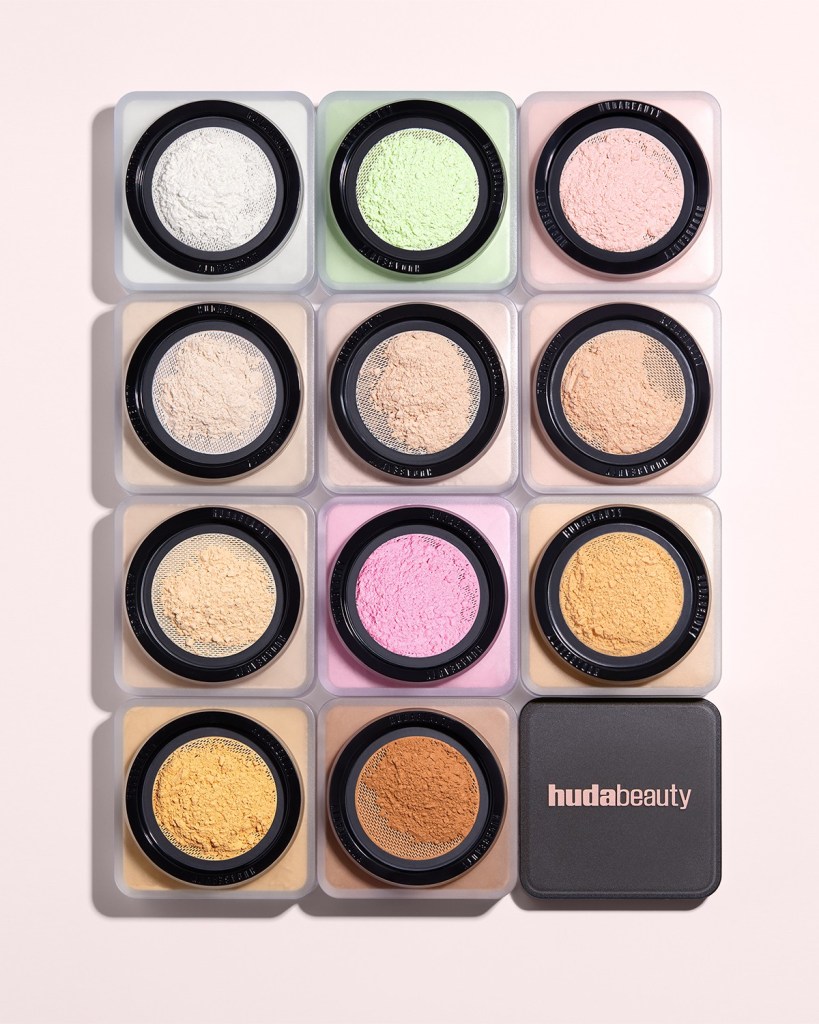 A flat lay arrangement of 12 containers of colored powders, including shades of white, green, pink, yellow, and beige, featuring a black circular mesh top. The containers are displayed on a light pink background, with a branded black compact labeled 'huda beauty' at the bottom right.