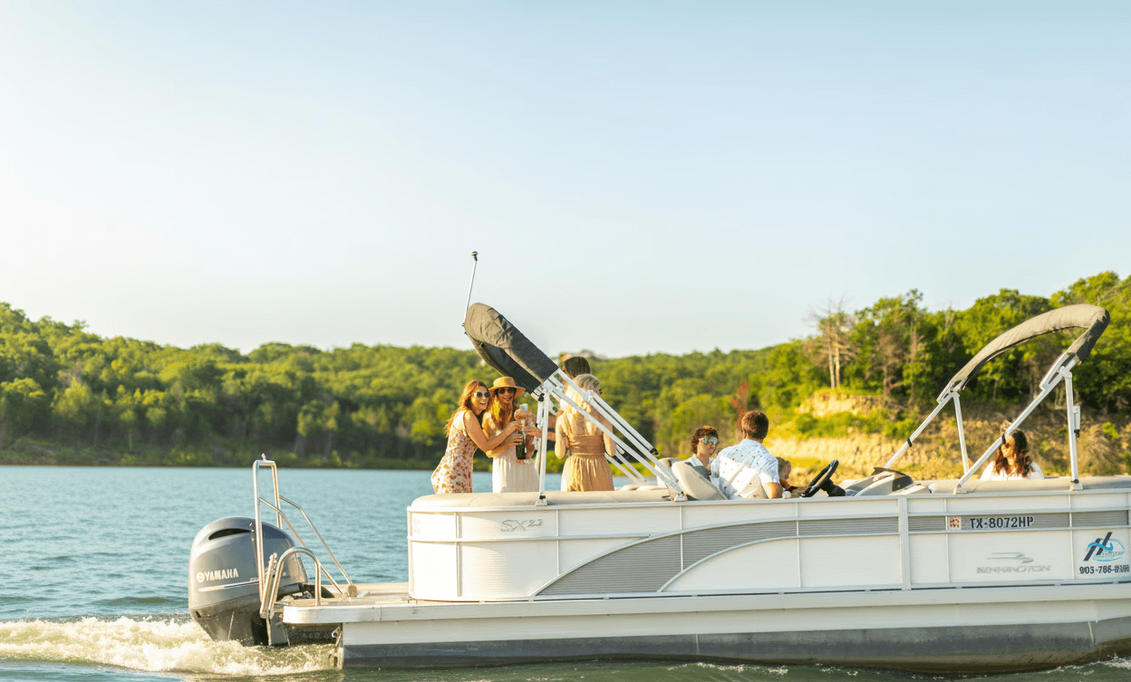 A group of people enjoy a sunny day on a stylish pontoon boat cruising across a serene lake, surrounded by lush green trees and gentle hills.