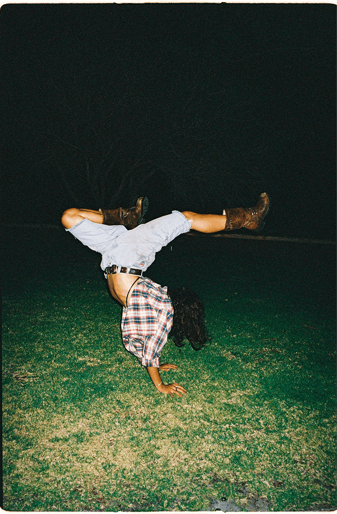 flash photo of a woman in a flannel shirt and denim shorts doing a cartwheel on grass at night
