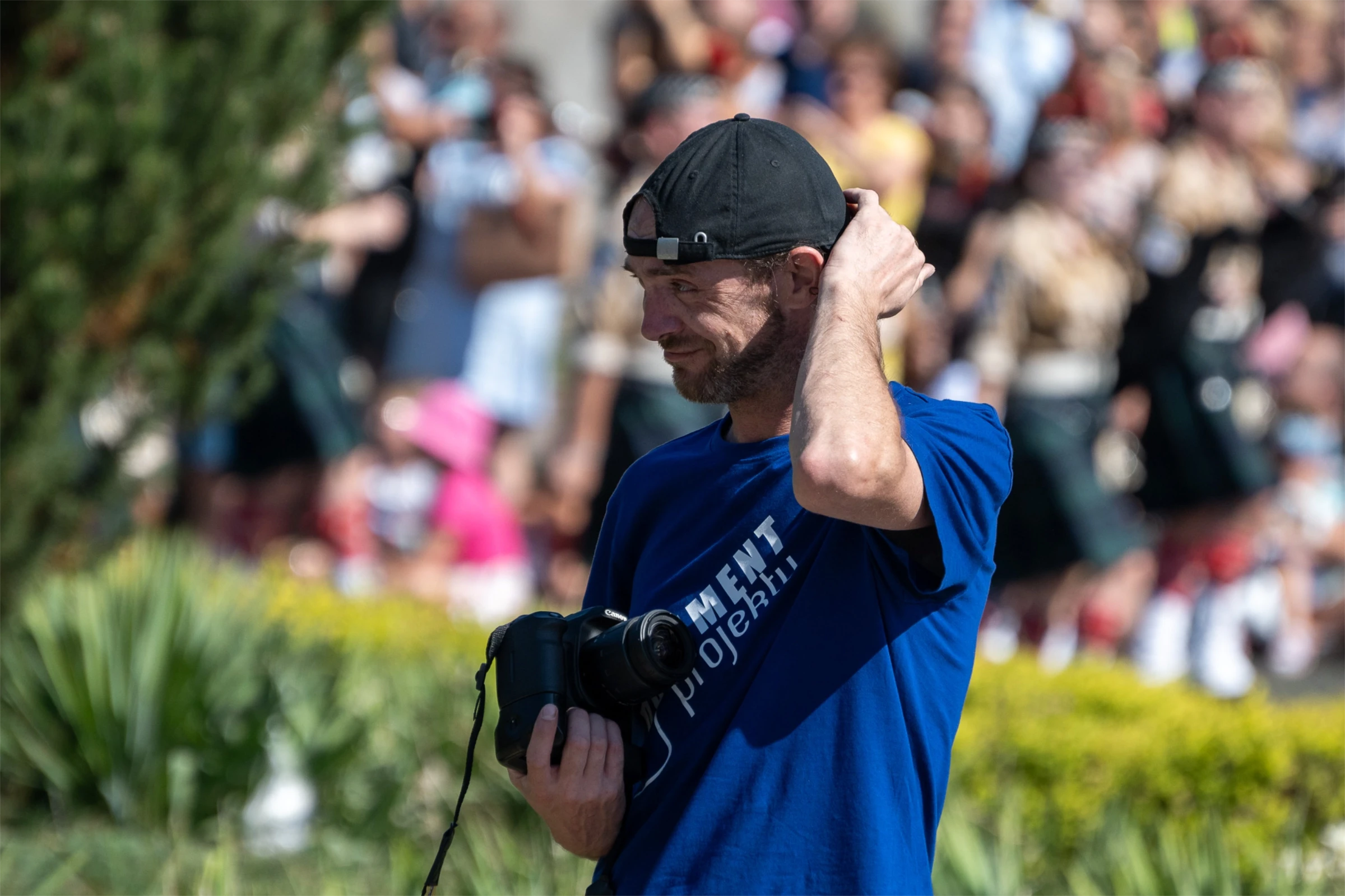 Mężczyzna w czapce z aparatem w ręku, poprawiający włosy. W tle ludzie i zieleń.