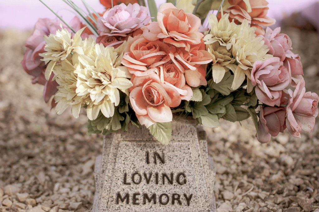 A bouquet of pastel-colored artificial flowers adorns a small granite gravestone engraved with "In Loving Memory," set against a backdrop of gravel and blurred greenery, symbolizing remembrance and honoring the departed.