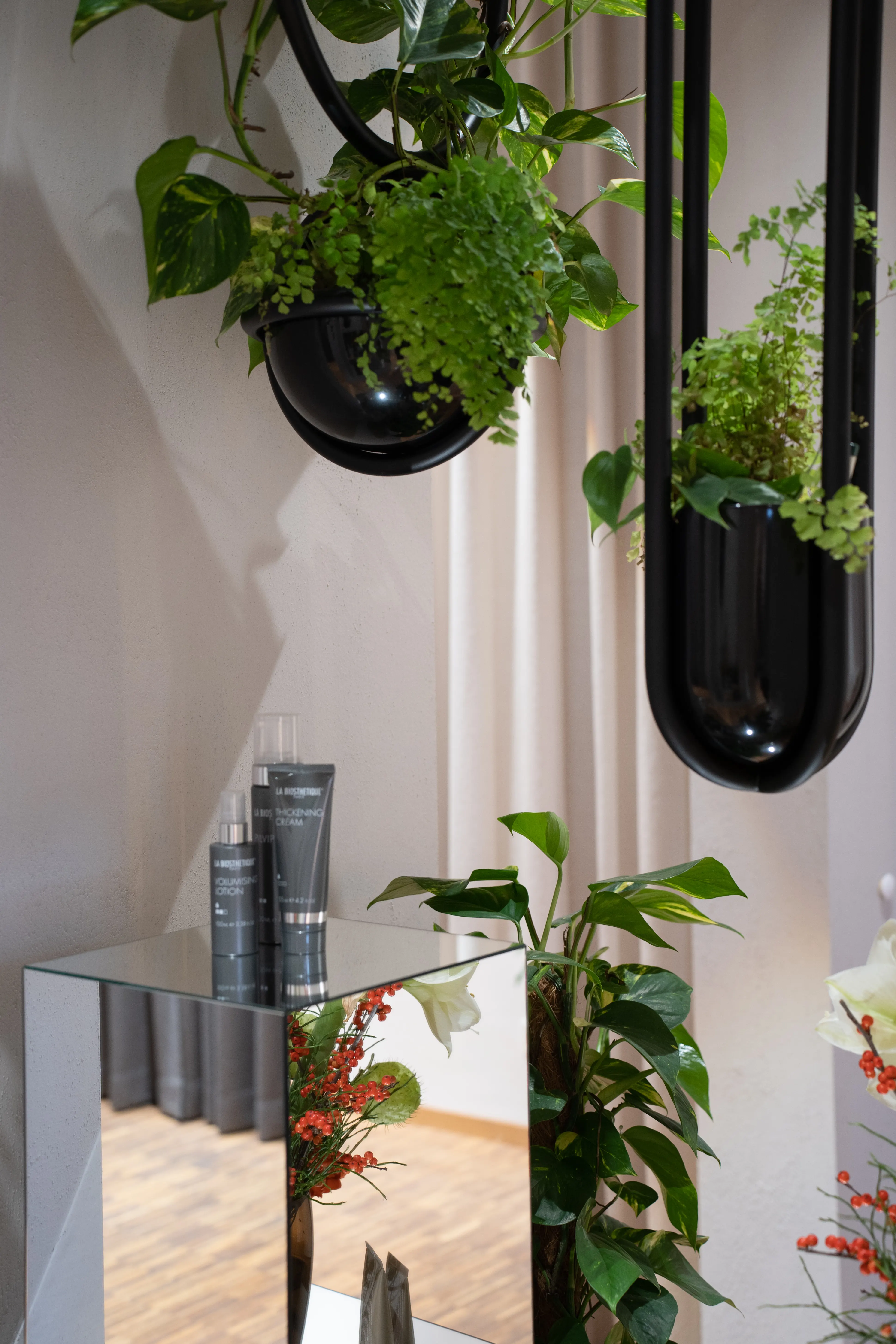 Detail of black oval hanging planters with ferns and trailing plants above mirrored cubes with hair products at EMPL FRISEURE.