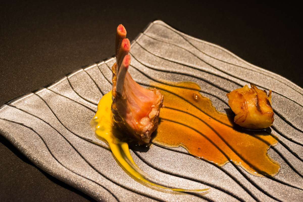 A piece of seared seafood with a curled tail sits on a striped napkin, drizzled with yellow and orange sauces.