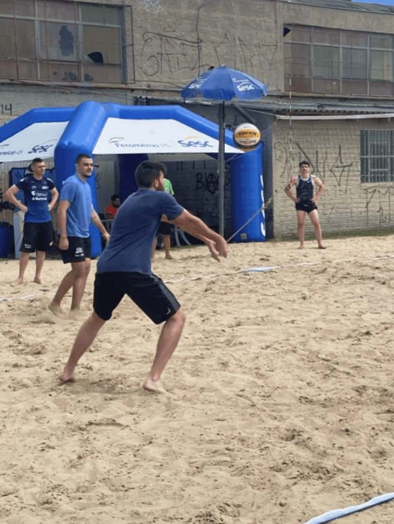 Augusto jogando vôlei de praia em uma quadra de areia, prestes a fazer um movimento para atingir a bola, com outras pessoas ao fundo e estruturas infláveis laterais.